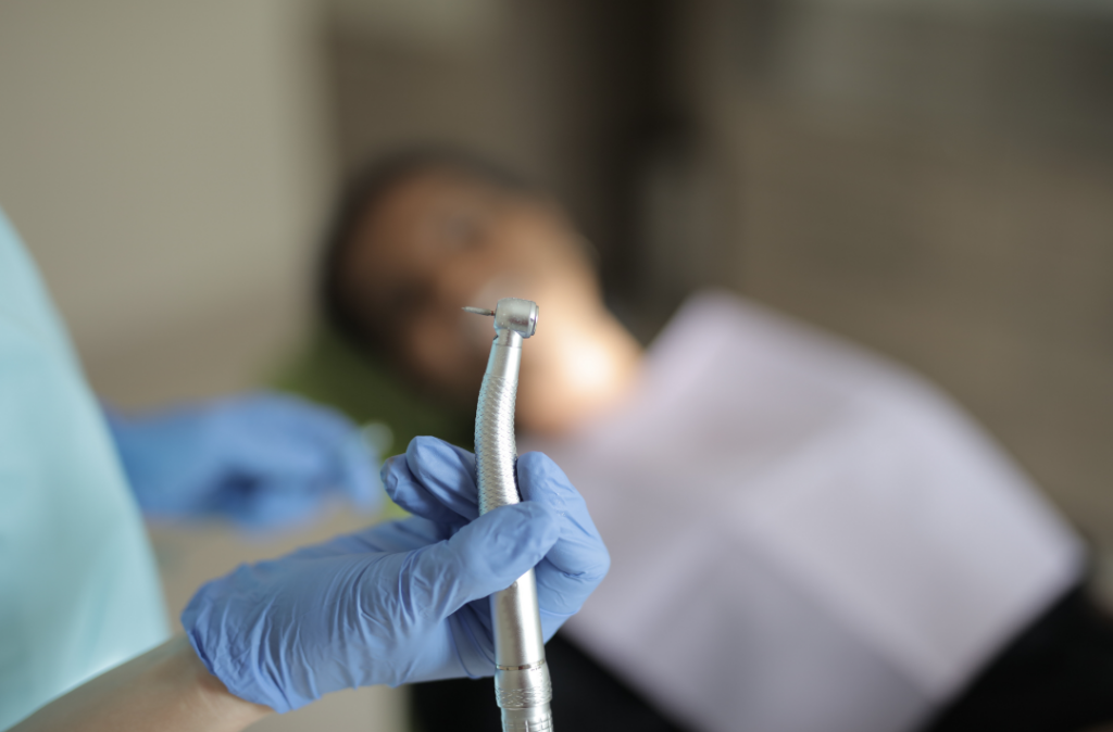 A gloved dentist holding a dental handpiece drill with a patient reclining in the background, illustrating dental anxiety triggers before a procedure.